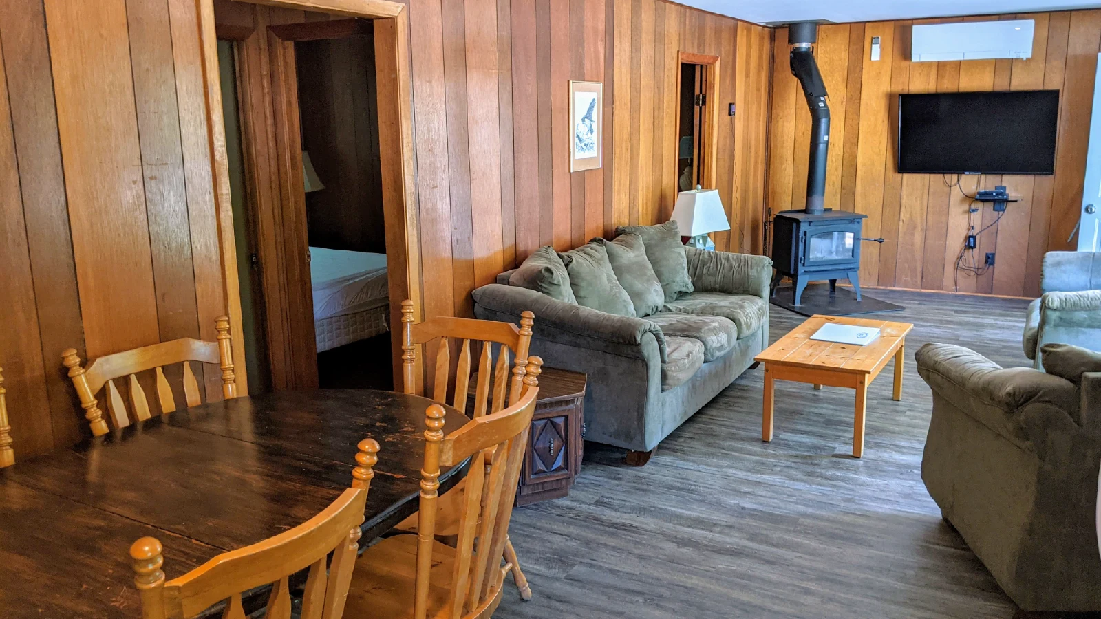 Pigeon Lake Resort &nbsp;Cottage nineteen.&nbsp; Open living and dining area with wood-paneled walls, table and chairs, sofa, wood stove, and television.